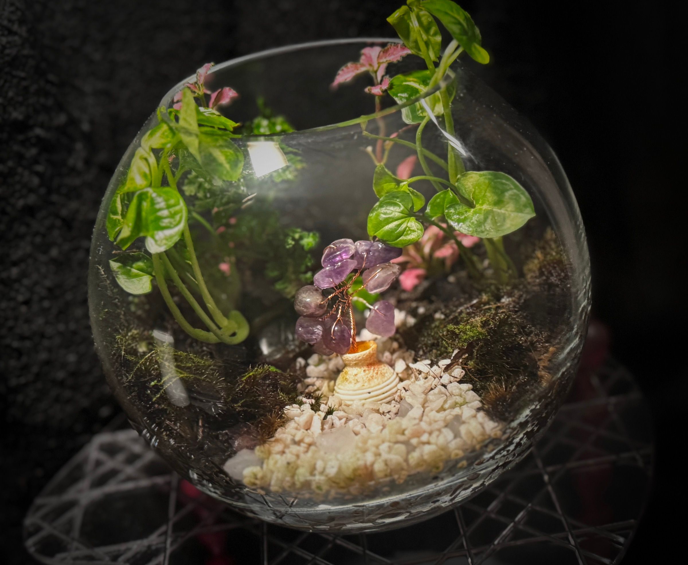 A glass globe vessel planted with moss and a wire amethyst tree over white gravel